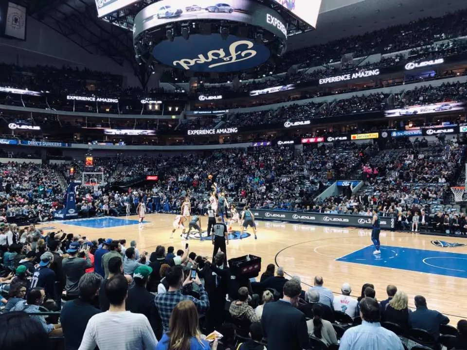 Live view of a Dallas Mavericks NBA basketball game at American Airlines Center, with players fiercely competing on the court and fans in the stands passionately cheering, immersing in the vibrant atmosphere.