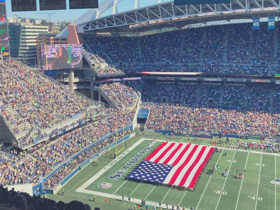 Panoramic view of Lumen Field stadium in Seattle, with a huge American flag covering the football field and stands packed with enthusiastic Seahawks fans ready for an NFL game.