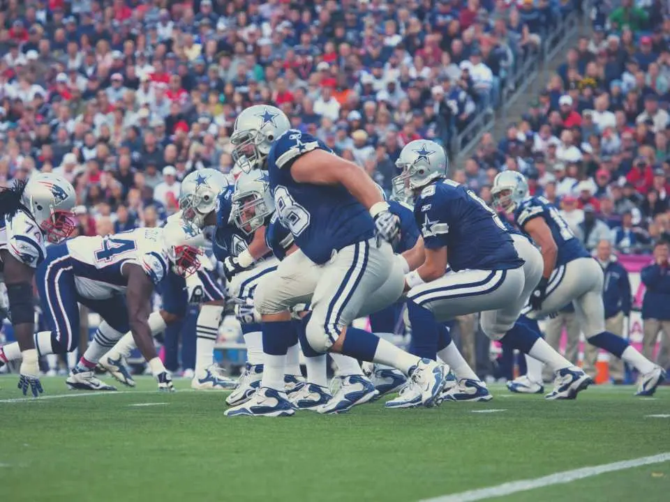 Dallas Cowboys football players in intense action on the field at AT&T Stadium, with passionate fans cheering in the packed stadium during an exciting NFL game.