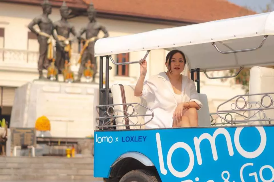 陽気な観光客がLOMO EVトゥクトゥクに乗り、背景には歴史的な三王の記念碑が見えます。チェンマイでの環境に優しい文化ツアーを楽しんでいます。