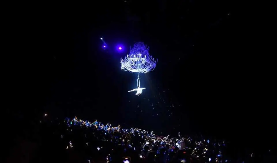 In the Shanghai Songcheng Show's aerial acrobatics, a stunt performer executes a thrilling act beneath a giant glowing chandelier, their silhouette prominent in the darkness, offering audiences a breathtaking experience.