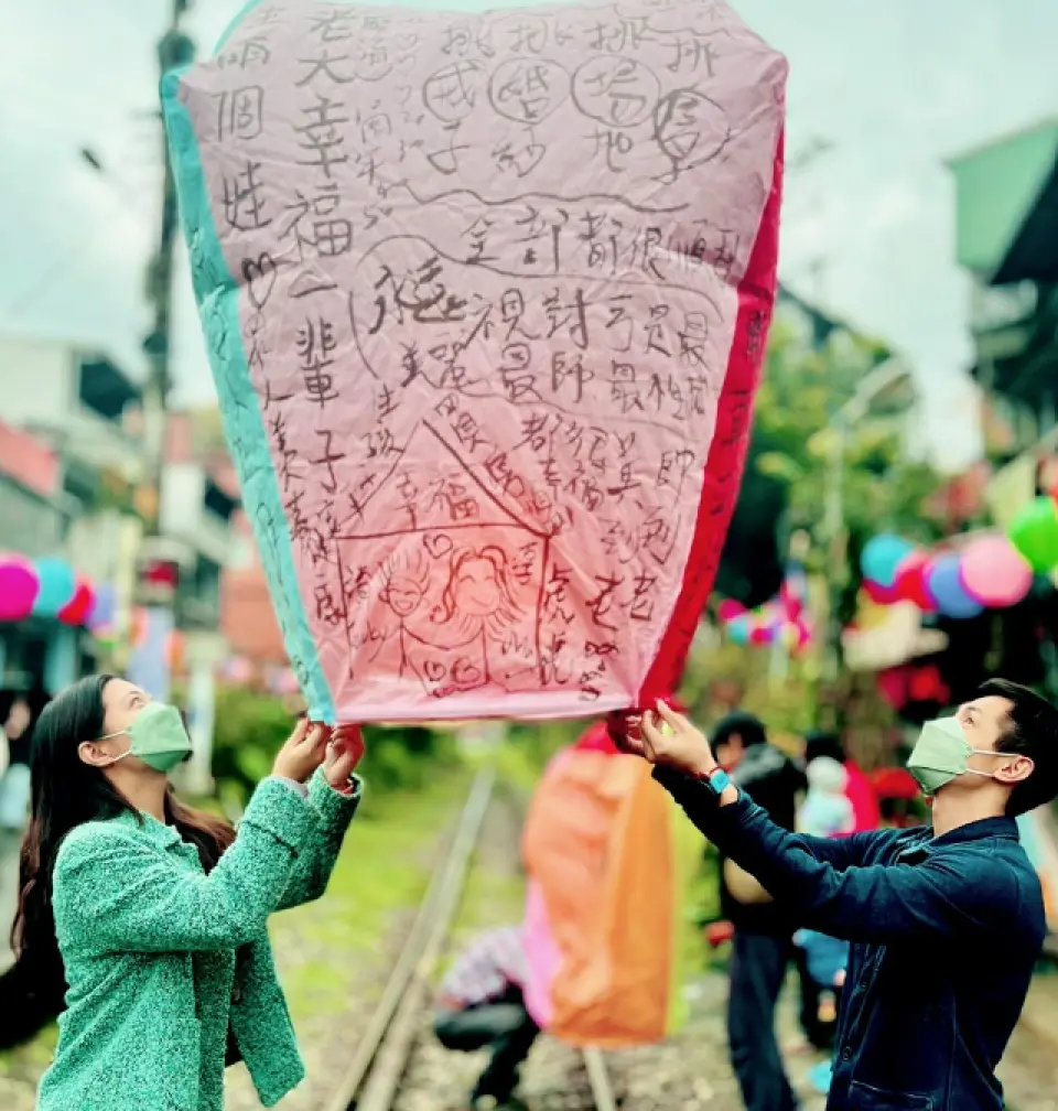 A couple wearing masks by the Pingxi railway tracks, focused on preparing to release a pink sky lantern filled with wishes, savoring Taiwan's traditional blessing culture.