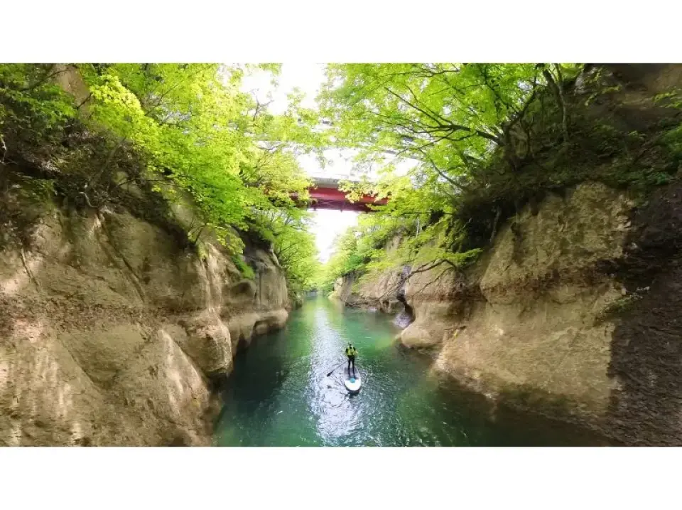 A traveler enjoys SUP paddling through the narrow canyon of Okura River Valley in Miyagi, Sendai, flanked by towering rock walls and lush green trees, with a red bridge visible in the distance, showcasing adventurous nature exploration.