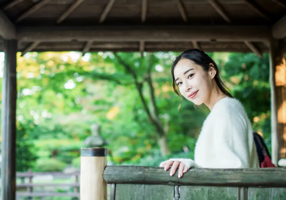 一位年輕女性在青森風景優美的日式亭閣中回眸微笑，展現優雅姿態，融入周圍的翠綠自然美景。