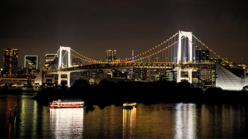 도쿄 레인보우 브릿지는 밤에 화려하게 빛나고, 멀리 도쿄 타워와 도시 야경이 선명하게 보이며, 그 아래로 유람선이 지나가며 도쿄만의 상징적인 랜드마크를 보여줍니다.