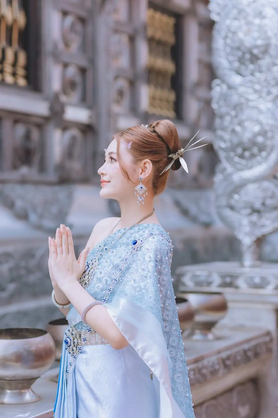 หญิงสาวในชุดไทยเชียงใหม่สวยงาม พนมมือไหว้ แสดงรายละเอียดเครื่องประดับผมและอัญมณีเด่นชัดท่ามกลางฉากหลังวัดศรีสุพรรณ