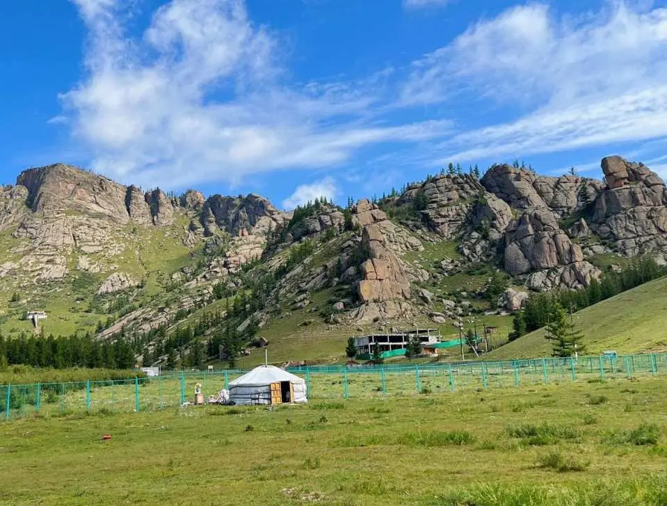 테를지 국립공원의 푸른 초원에 자리한 전통 몽골 게르, 웅장한 바위산과 맑은 푸른 하늘을 배경으로, 특별한 숙박 경험을 선사합니다.