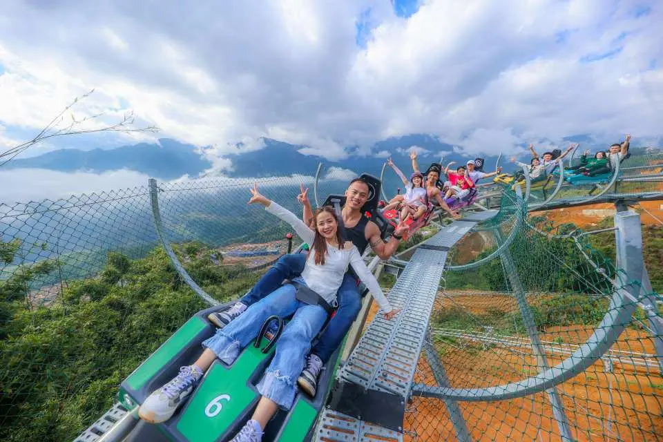 ผู้เยี่ยมชมโบกมืออย่างมีความสุขจากรถรางบนรถไฟเหาะอัลไพน์ ซาปา บานมอง เพลิดเพลินกับความตื่นเต้นของการขับขี่ผ่านเทือกเขาฮว่างเลียนเซินและมองเห็นหุบเขา.