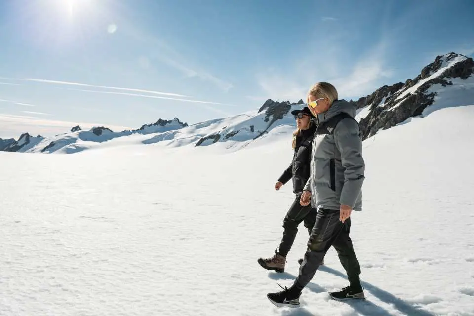 Two visitors on New Zealand's Mount Cook Ultimate Alpine Air Tour, walking on the sunny Tasman Glacier, experiencing the stunning snow-covered Southern Alps landscape.