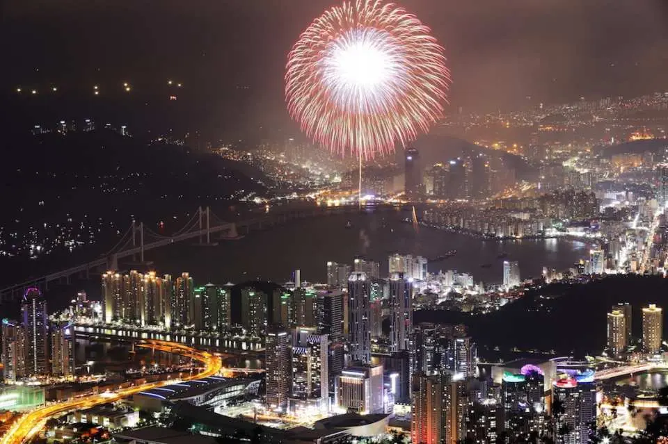 釜山煙火節單一巨型煙火在高空璀璨綻放，俯瞰下方的釜山城市天際線與燈火通明的橋樑。