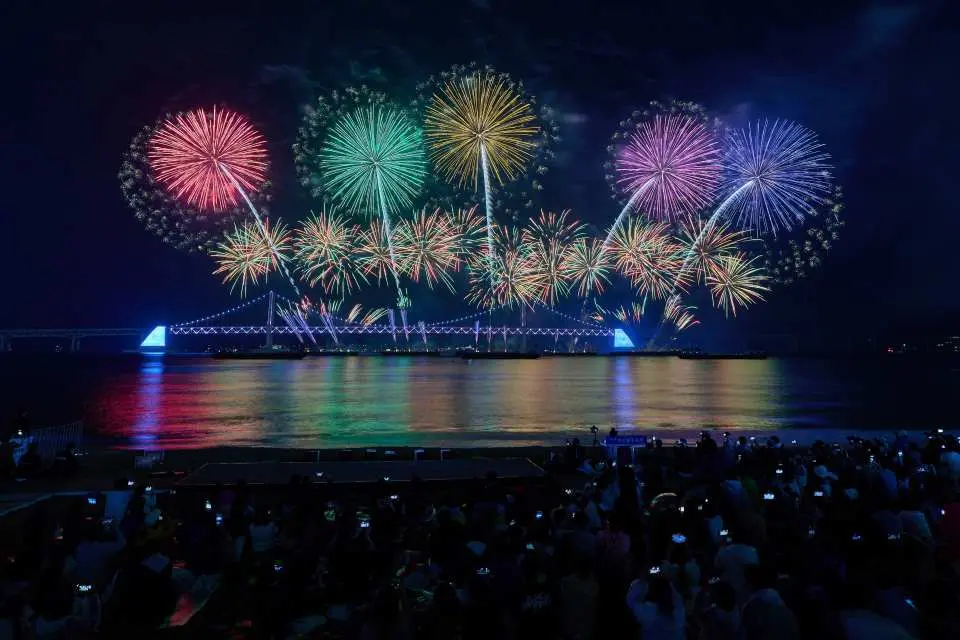廣安里海水浴場的觀眾聚集欣賞2025釜山煙火節，廣安大橋上方的絢爛煙火與雷射燈光秀倒映海面。