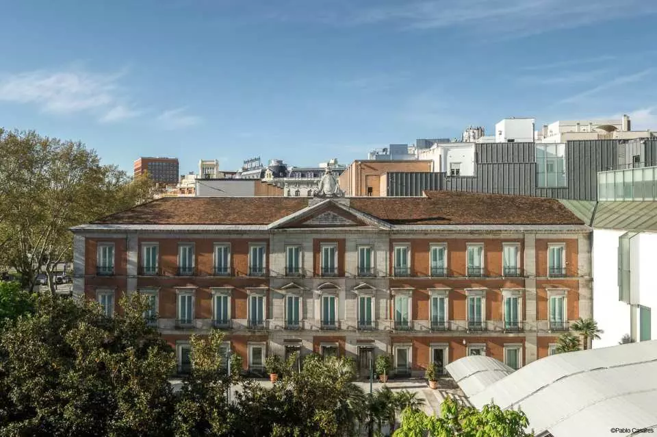 The elegant facade of a classical Madrid building, possibly a museum or art gallery, with rows of windows and a red-tiled roof, surrounded by lush green trees under a clear blue sky.