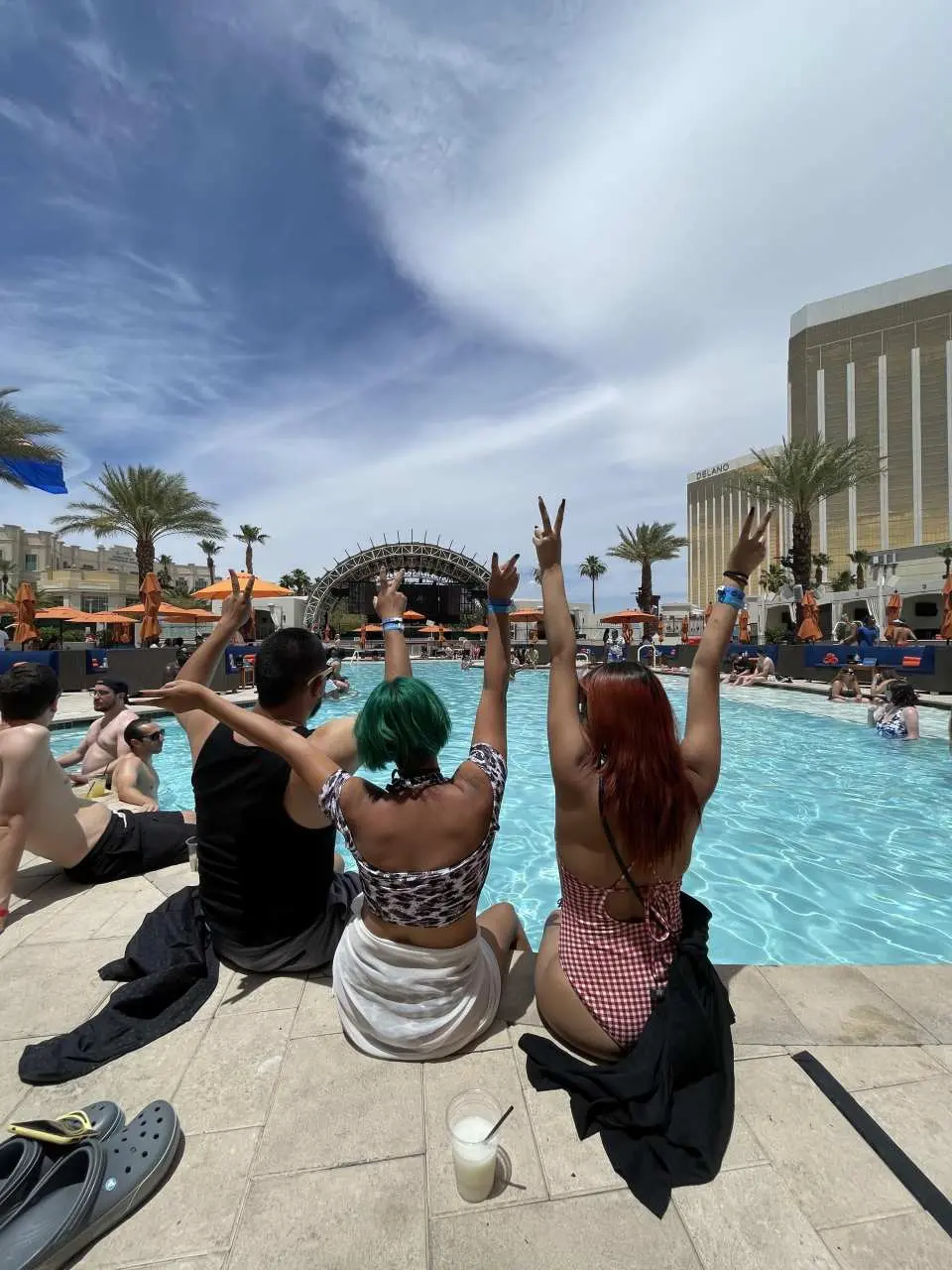 Friends enjoying a vibrant day club pool party in Las Vegas, raising peace signs by the poolside with a bustling resort background, showcasing the ultimate party experience.