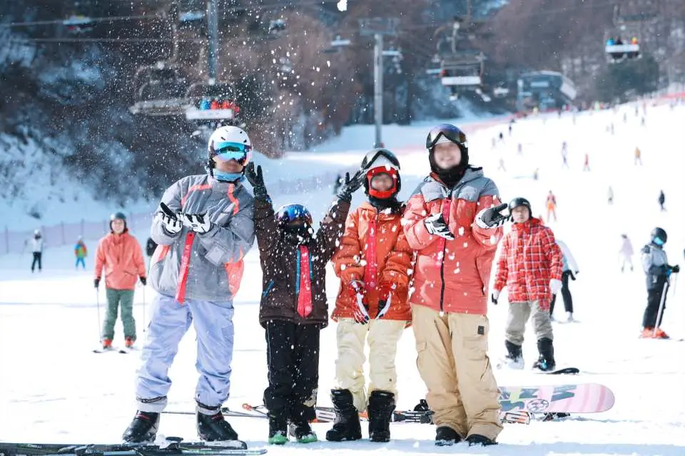A family or group of friends happily poses for a photo at Elysian Gangchon Ski Resort as snow falls, with a busy ski slope and lifts in the background, showing winter skiing's warmth and vibrancy.