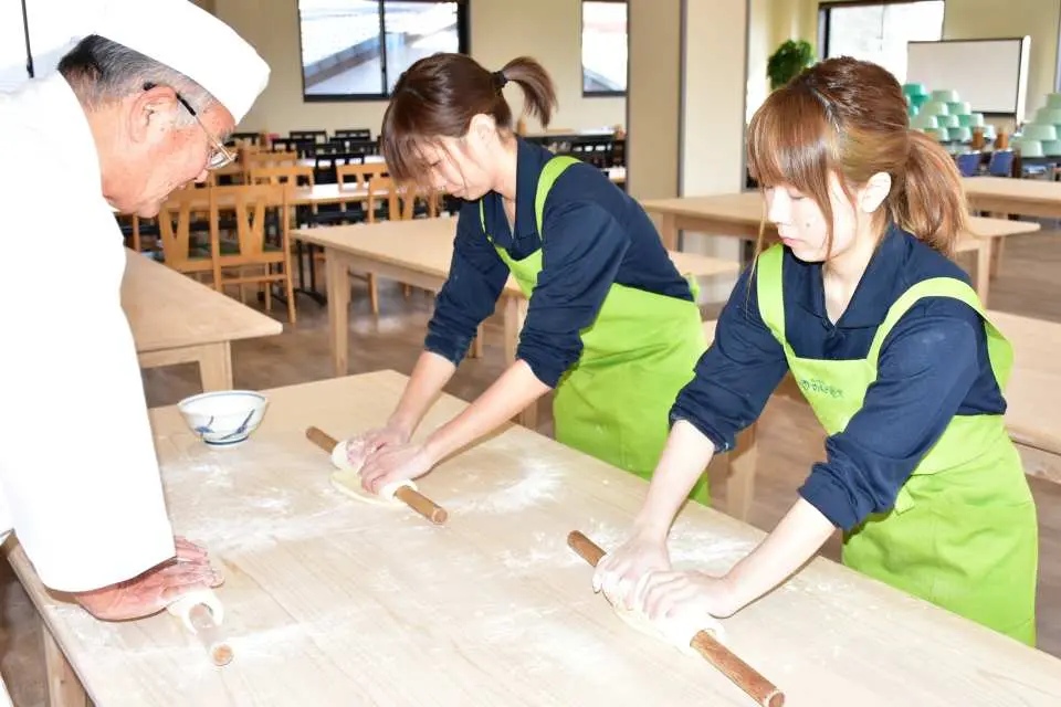 高松市の讃岐うどん作り体験教室で、2人の参加者が集中して麺棒でうどん生地を伸ばしており、うどんマスターが指導しています。