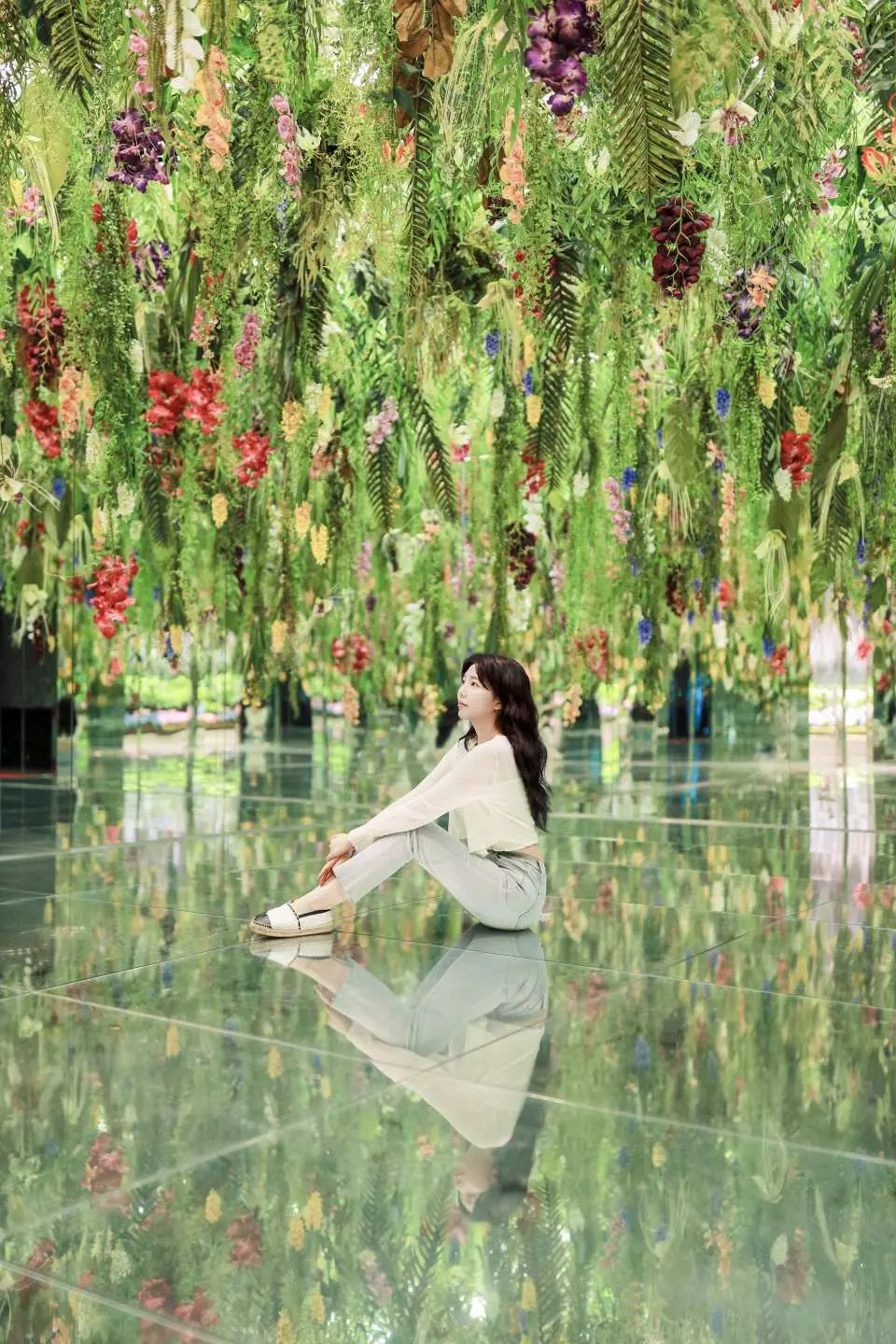 A woman elegantly sits on the mirrored floor of Gapyeong Begonia Bird Garden's Mysterious Garden, enjoying the magical atmosphere surrounded by reflected flowers and plants.