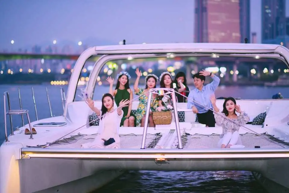 A group of young friends happily waving on the deck of a luxury yacht on Seoul's Han River at dusk, enjoying a joyful yacht experience with city lights in the background.