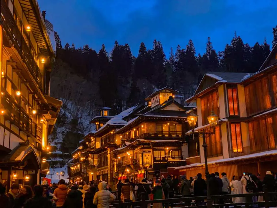Bustling night view of Ginzan Onsen in Yamagata, Tohoku, showcasing elegantly lit Taisho-era buildings and snowy streets filled with visitors enjoying this magical hot spring town.