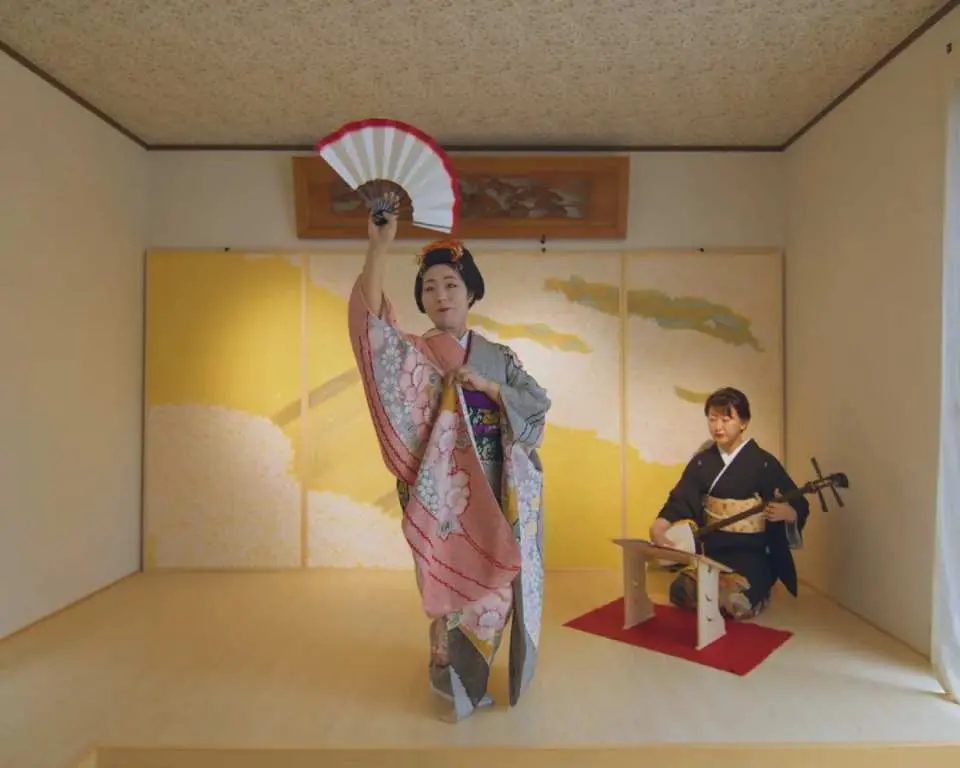 An elegant kimono dancer performing a traditional Japanese dance with a fan in a Hakata setting, accompanied by a shamisen player, showcasing rich Japanese culture.