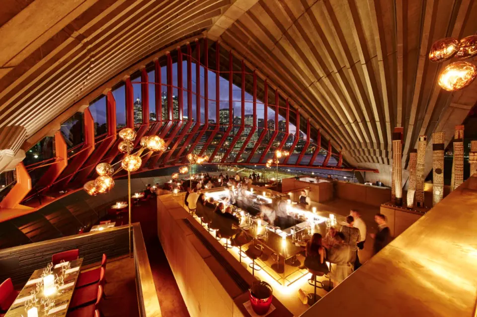 Interior of Bennelong restaurant at Sydney Opera House, showcasing elegant dining and bar with panoramic views of the illuminated Sydney skyline at dusk, offering a fine Australian dining experience.