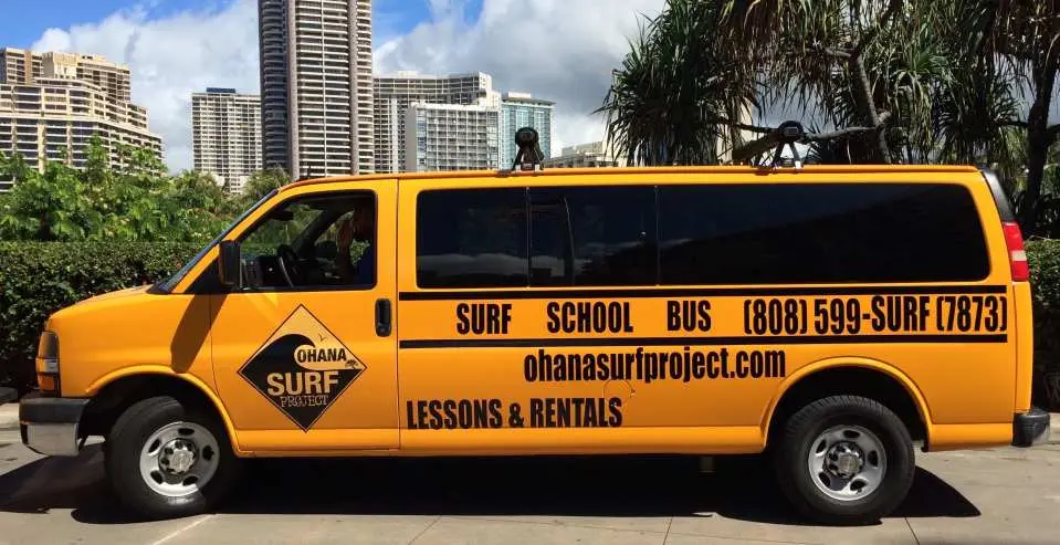 The yellow "OHANA SURF PROJECT" surf school bus offering convenient Waikiki hotel transfers, clearly branded with contact information, ready to transport students.