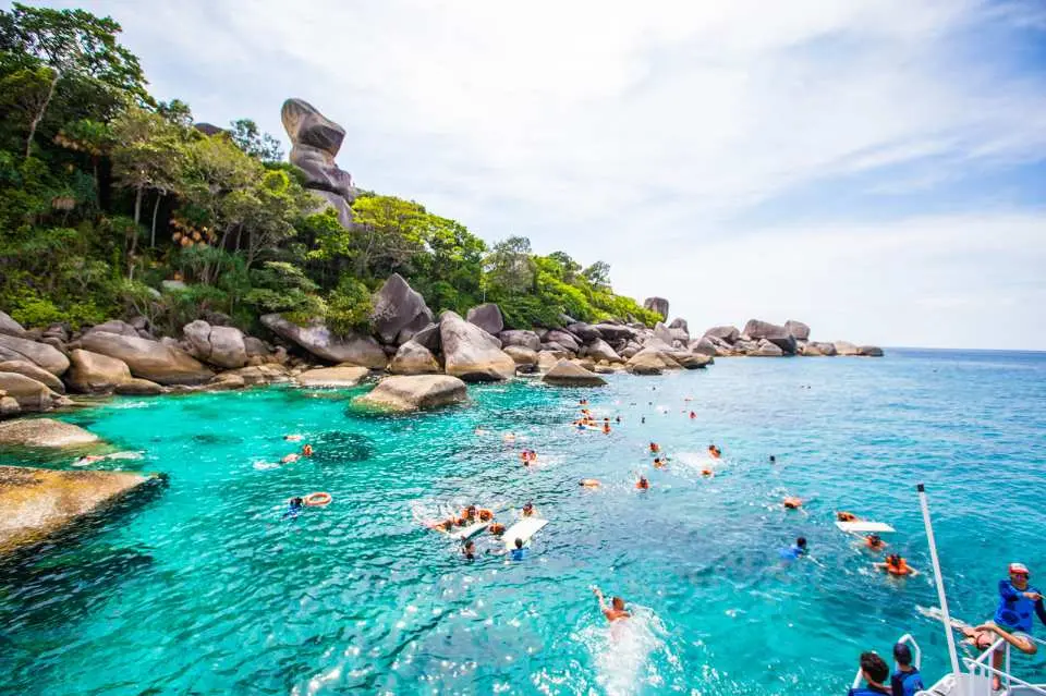 Tourists enthusiastically snorkeling and swimming in the clear turquoise waters near the rocky coastline of Similan Islands, exploring colorful coral reefs during a speedboat snorkeling trip from Khao Lak.