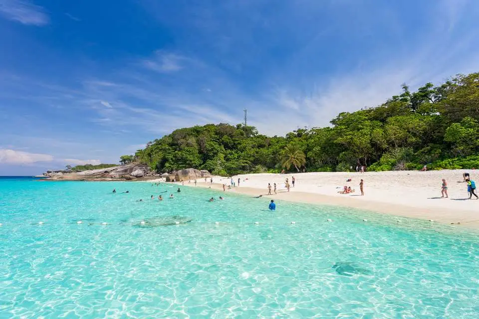 A long, pristine white sand beach at Similan Islands, bustling with tourists enjoying the sun, clear blue sky, and turquoise waters, backed by dense tropical jungle, offering an ideal island getaway from Khao Lak.
