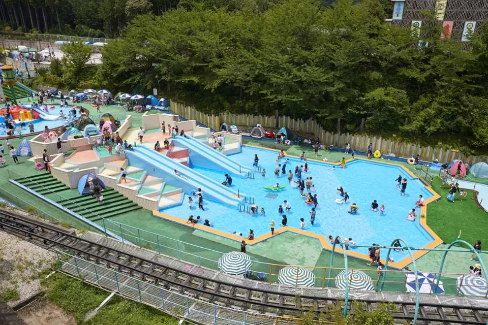 大阪の関西サイクルスポーツセンター水上公園の全景。2つの大きな青いウォータースライダーがあり、多くの訪問者が太陽が降り注ぐプールで楽しんでいます。