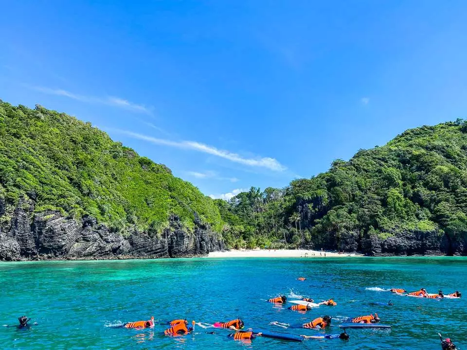 Tourists in snorkeling gear explore the vibrant underwater world in the clear turquoise waters of Thailand's Maya Bay or Phi Phi Islands, enjoying close encounters with marine life.