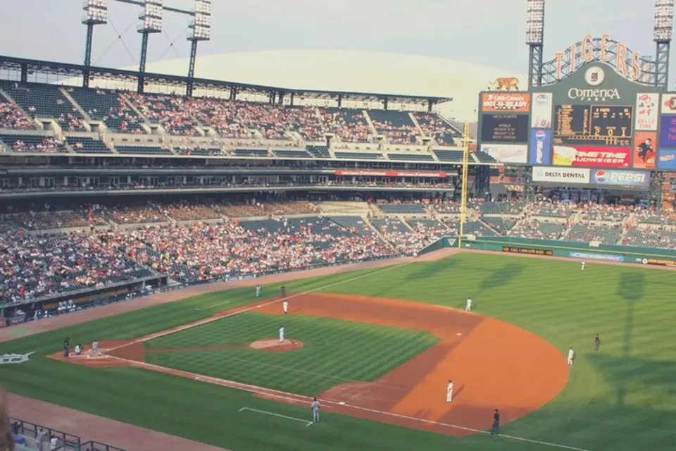 Daytime panoramic view of Comerica Park baseball stadium during an MLB Detroit Tigers game, showcasing the lush green field, full stands of cheering fans, and the iconic scoreboard.