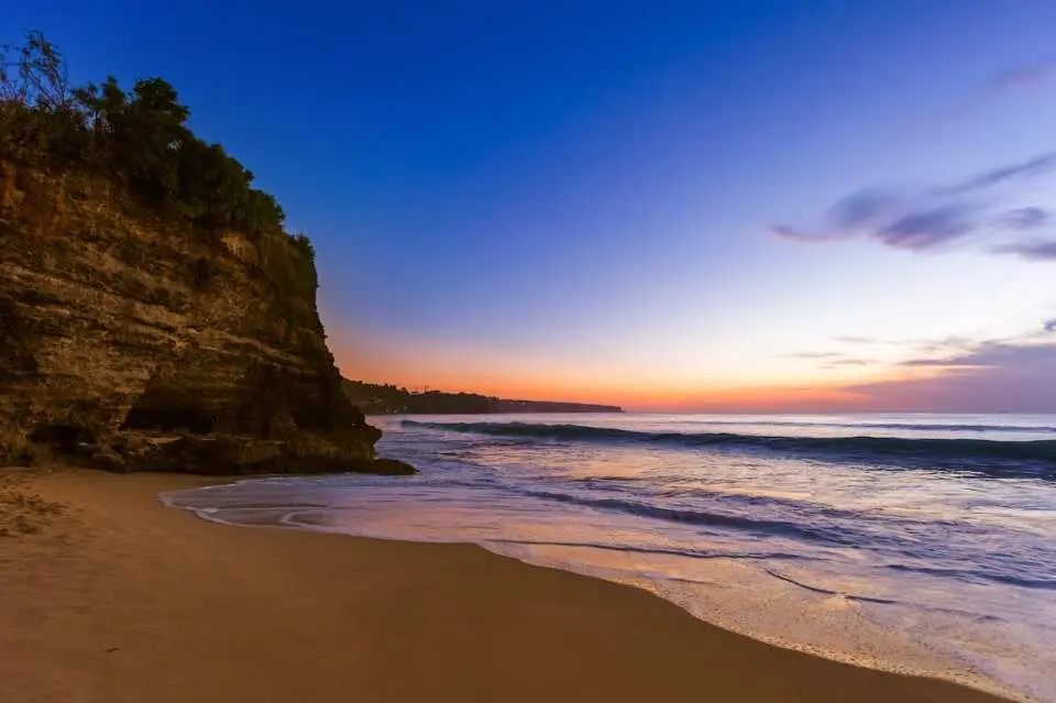 Tranquil twilight at Uluwatu Dreamland Beach, gentle waves lapping golden sand, with soft colors painting the distant horizon, embracing Bali's island romance.