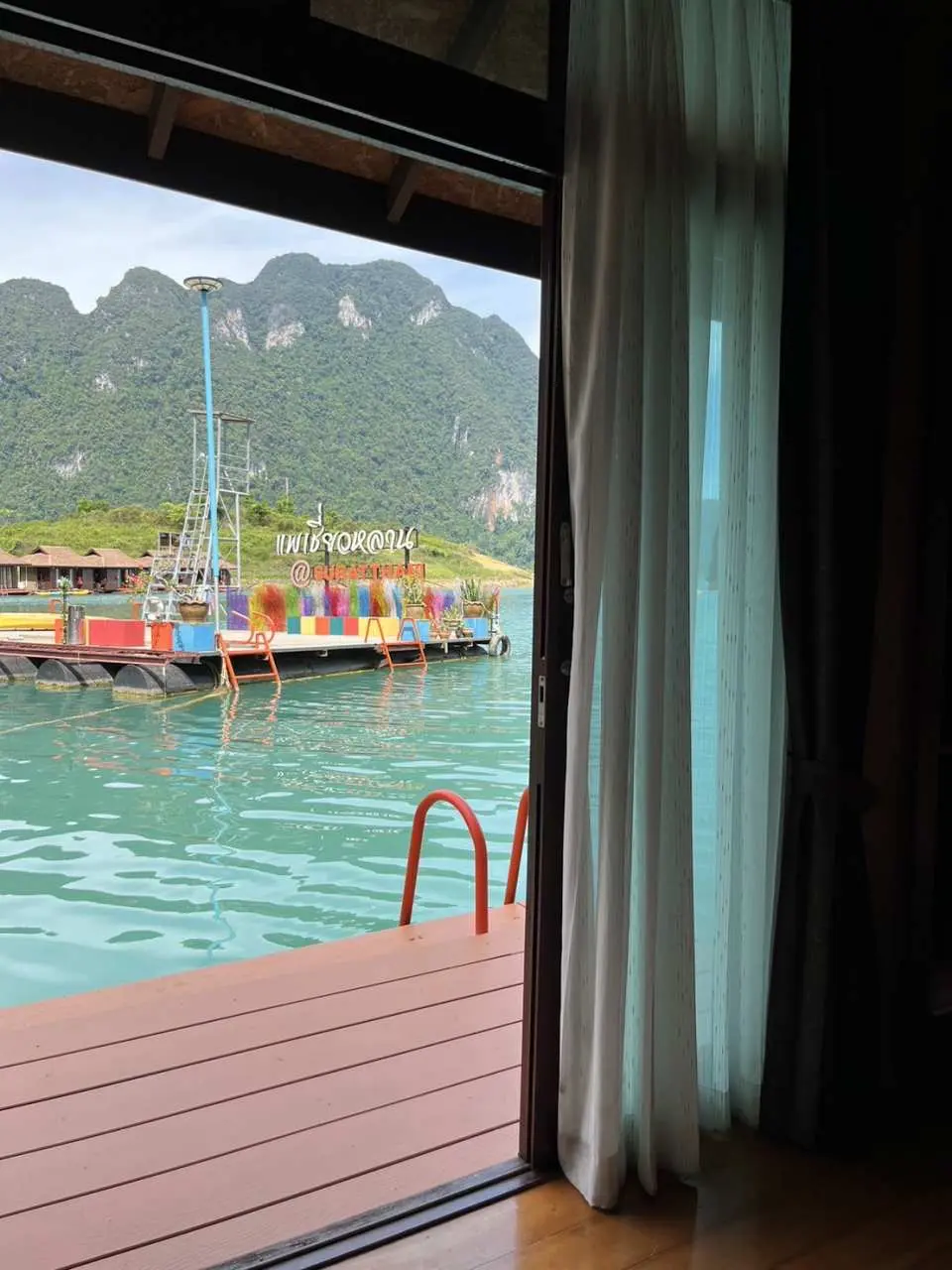 View from a Khao Sok Lake floating bamboo bungalow window, showing a colorful dock with boats moored, and majestic limestone mountains in the distance, showcasing the unique scenery of a Thai national park.