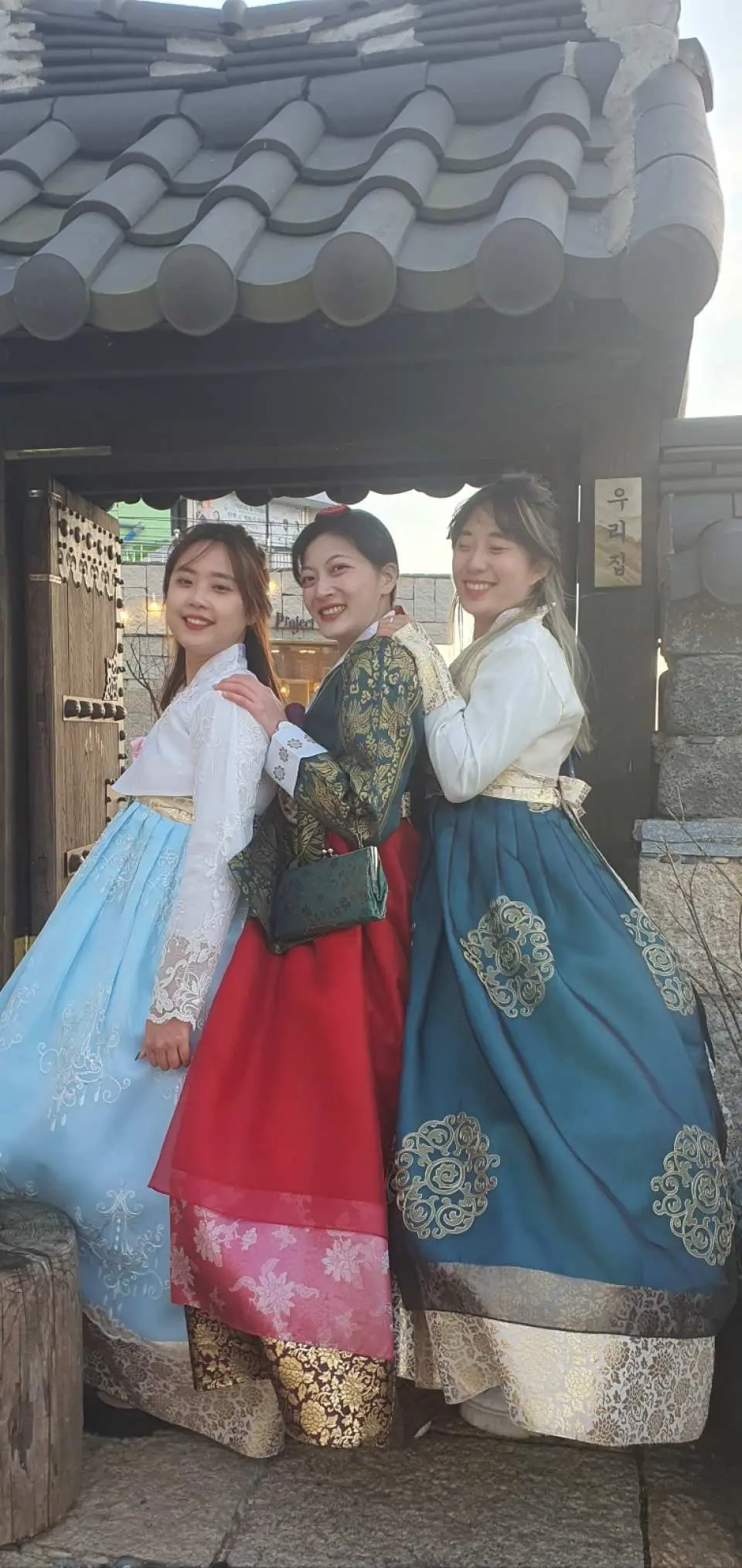 三位面帶笑容的女子，分別穿著淺藍、紅色和青綠韓服，展示多樣風格，在韓國傳統瓦片屋頂背景下親密合影。