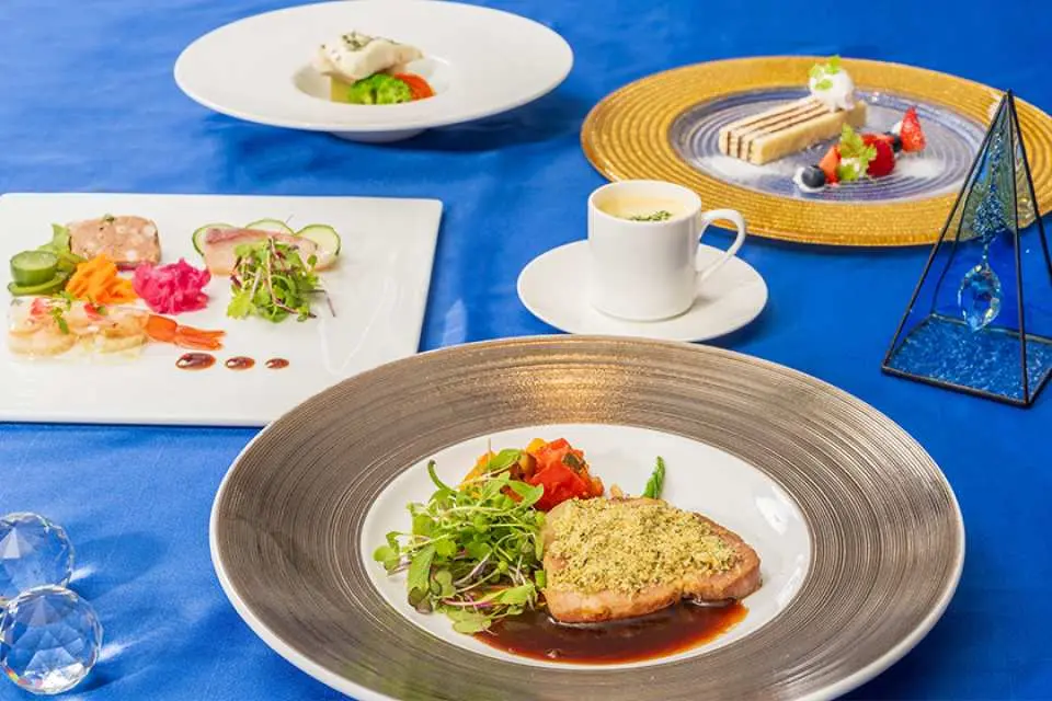 神戸クルーズ船の青いテーブルクロスに並べられた芸術的な洋食フルコース。メイン料理、多様な前菜、美しいデザートがロマンチックな食事の雰囲気を演出。