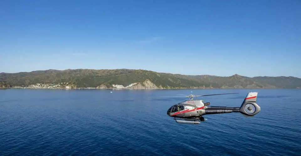 直升機在加州卡塔利娜島湛藍海面上空飛行，遠方連綿起伏的山脈勾勒出壯麗海岸線，享受空中遊覽。