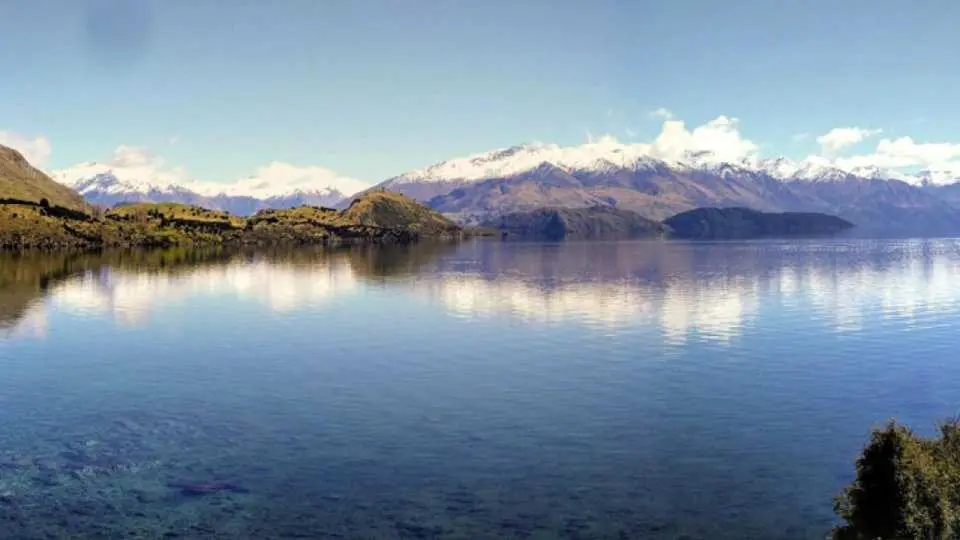 Panoramic view of Lake Wanaka's crystal-clear waters, reflecting distant snow-capped mountains, showcasing New Zealand's pristine natural beauty surrounding Ruby Island.