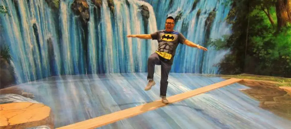 一名身穿蝙蝠侠T恤的男子，在芭提雅3D美术馆的瀑布错视画作中，沿着木板摆出平衡姿势，展现惊险又有趣的互动拍照体验。