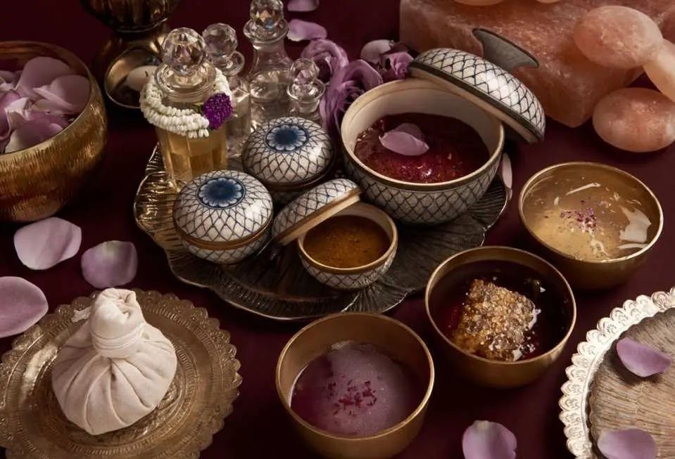 曼谷Divana Scentuara Spa泰式水療產品近景，展示多種天然磨砂膏、精油和傳統草藥球，搭配紫色花瓣，營造典雅氣氛。