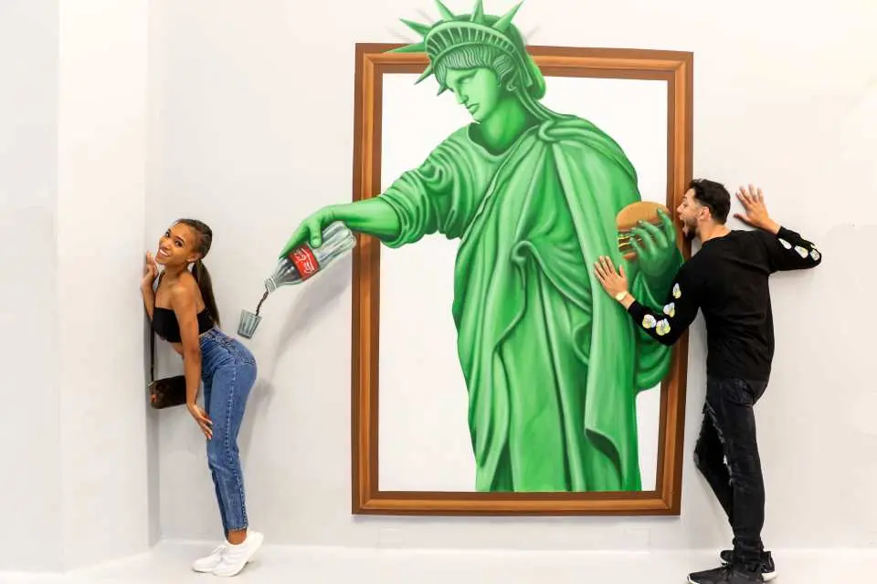 Visitors engaging with a playful 3D illusion of the Statue of Liberty pouring soda with a fork and a person offering a hamburger, creating humorous and perception-challenging photo opportunities at the Los Angeles Museum of Illusions.