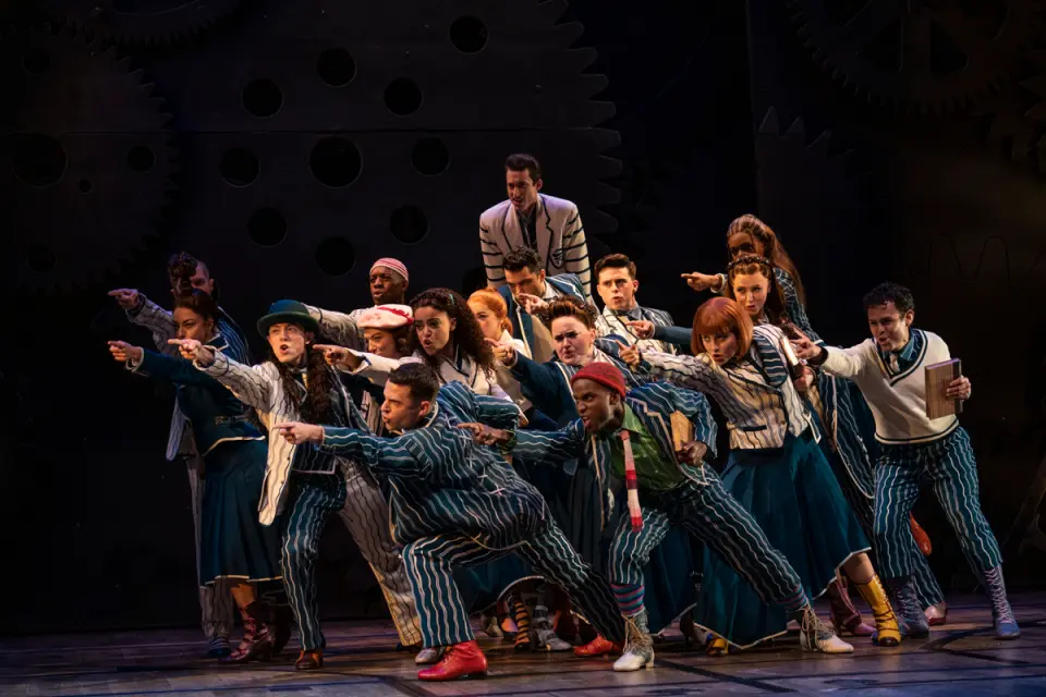 紐約百老匯《Wicked》音樂劇中一群演員穿著條紋服裝，動作一致地指向前方，充滿活力。