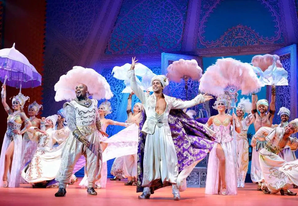 百老匯《阿拉丁》音樂劇中，身穿銀白色華麗戲服的舞者們，手持巨型羽毛扇和花朵造型道具，在舞台上表演，充滿魔幻。