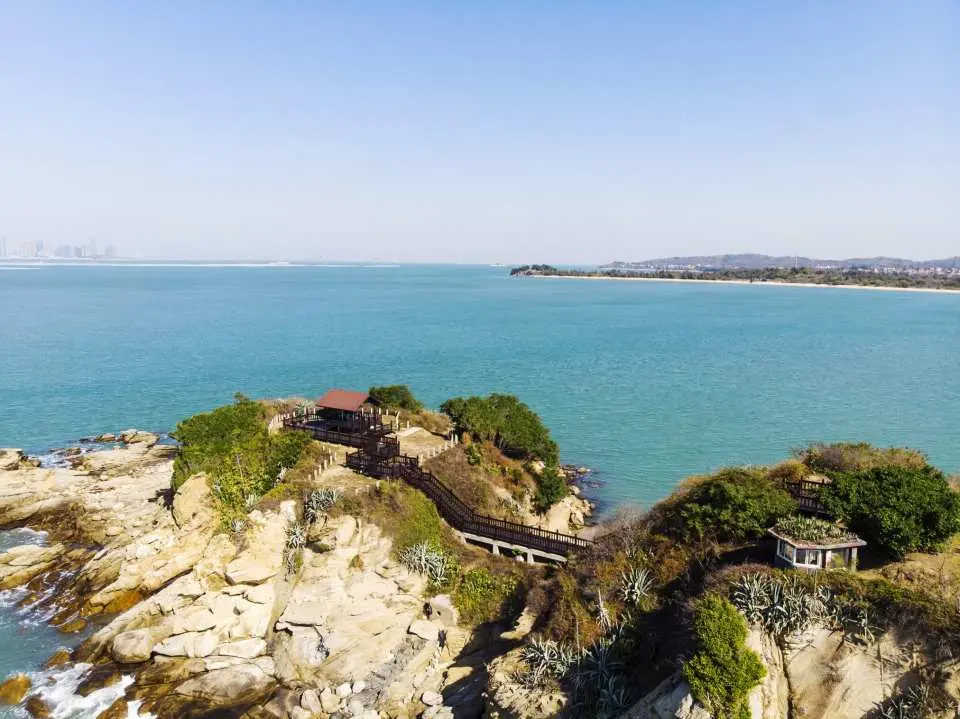 金門烈嶼沙溪堡空拍全景，碧海藍天與崎嶇岩岸構成壯麗景色，遠處可見金門本島，展現小金門自然之美。