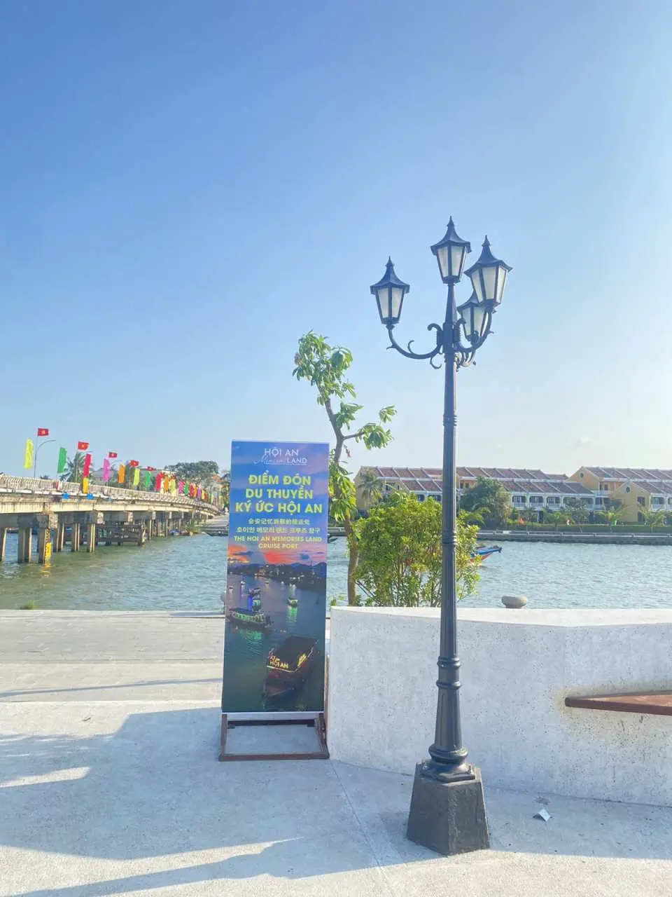 會安印象主題公園入口處的廣告牌與古典路燈，背景是寧靜的河流與遠方的橋樑，晴朗天氣下的會安景緻。