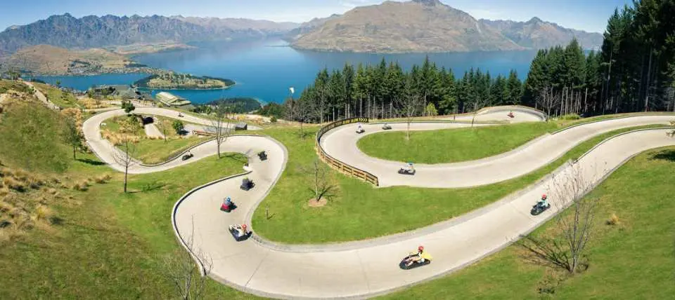 Adventurous guests race down a winding luge track at Skyline Queenstown, enjoying thrilling fun with stunning views of Lake Wakatipu and green hills.