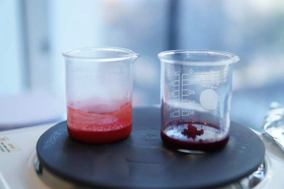 A demonstration in a Korean beauty DIY class, showing the ingredient mixing process for making lipsticks or skincare products, with two beakers of different red liquid concentrations being heated.