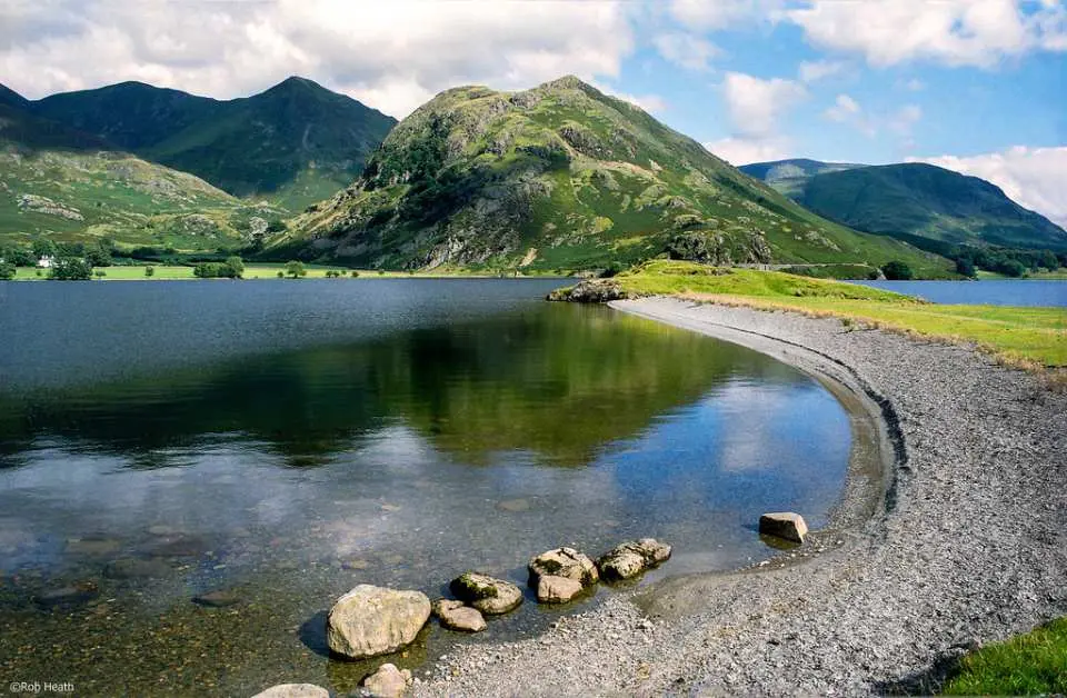 A picturesque view of Windermere Lake in the Lake District, England, with tranquil waters reflecting green mountains and a cloudy sky, showcasing the region's natural beauty.