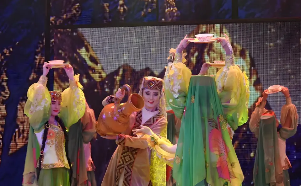 An ensemble performance from Xinjiang's 'A Thousand Times West Region' show. Dancers in vibrant green ethnic costumes balance bowls and hold traditional pitchers, showcasing acrobatic skills and diverse Western Regions culture.