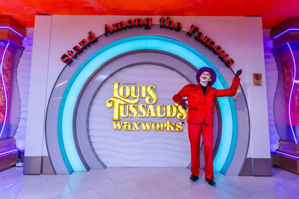 芭堤雅路易斯杜莎夫人蠟像館入口，一位穿紅色西裝的逼真蠟像擺出姿勢，背景是品牌標誌和時尚弧形設計，歡迎遊客與名人合影。