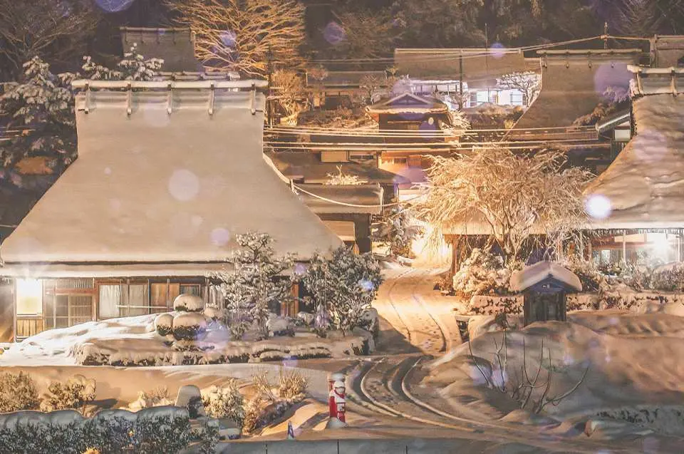 京都美山かやぶきの里の冬の夜。伝統的な茅葺き屋根の家々が厚い雪に覆われ、暖かい光が屋根の輪郭を照らし出し、静かで魅力的な雪国の夜景を作り出しています。