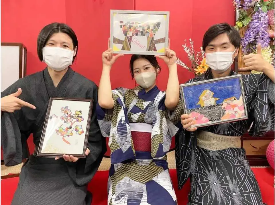 Three smiling participants in traditional Japanese kimonos proudly presenting their finished, framed Kimono Hagi art creations after a creative indoor experience in Kanazawa.